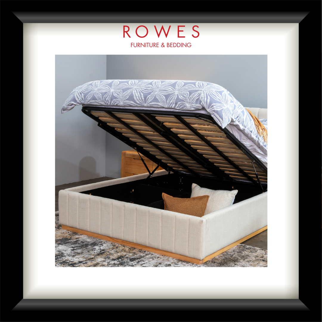 A bedroom scene featuring a large, plush bed with a fabric bedhead and foot, bedsides and a large rug. The bed is up and open, demonstrating the storage hidden underneath the bed. The Rowes Furniture logo features