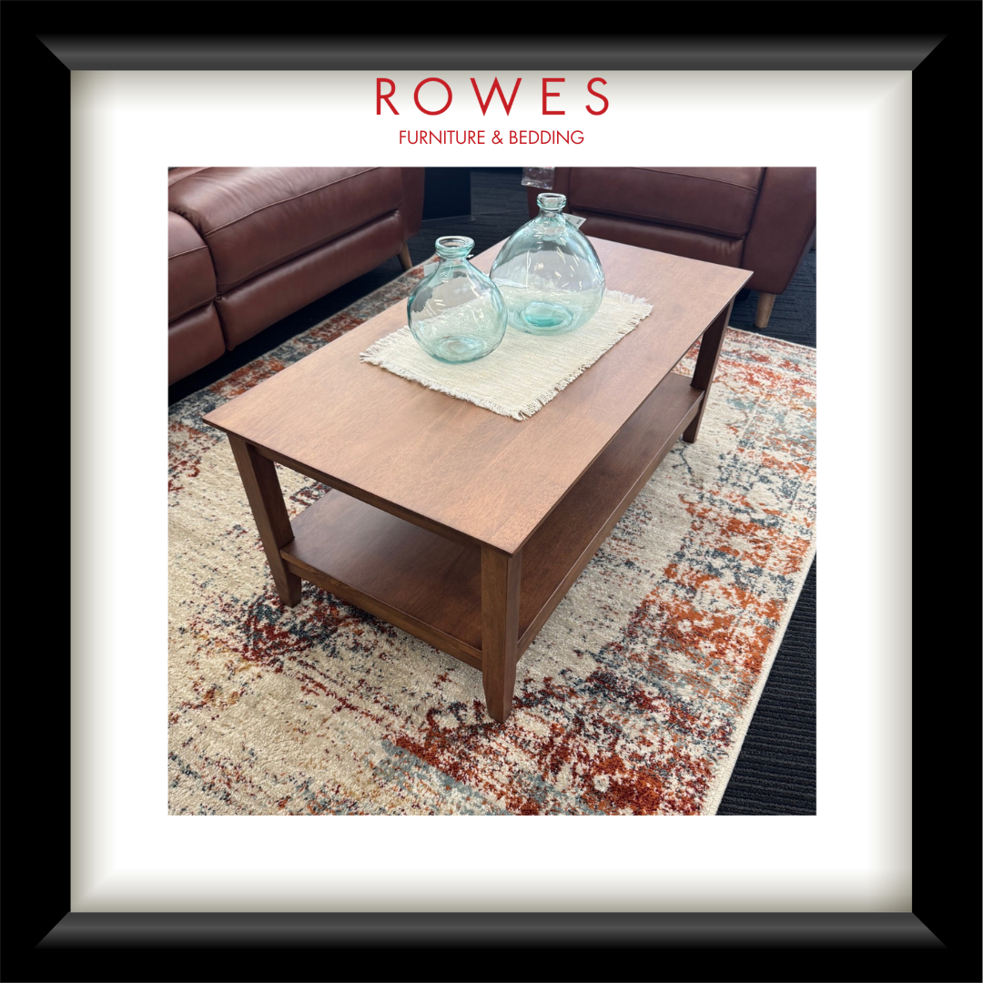 A framed interior photo print showing a wooden coffee table in a living room setting with two clear glass bottles on top, placed on a patterned rug. The print is matted with a white border and enclosed in a black frame.