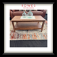 A framed interior photo print showing a wooden coffee table in a living room setting with two clear glass bottles on top, placed on a patterned rug. The print is matted with a white border and enclosed in a black frame.