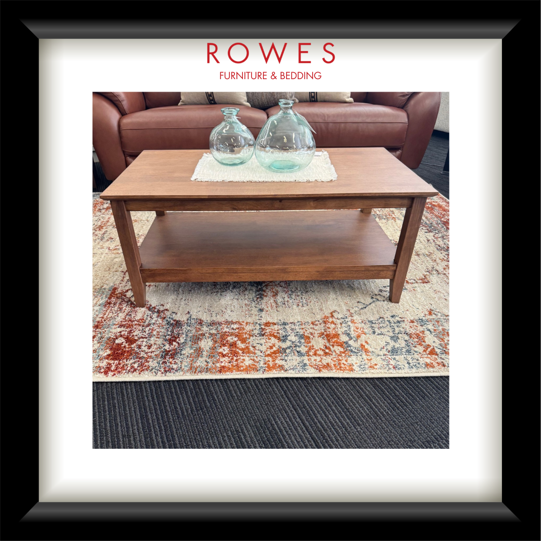 A framed interior photo print showing a wooden coffee table in a living room setting with two clear glass bottles on top, placed on a patterned rug. The print is matted with a white border and enclosed in a black frame.
